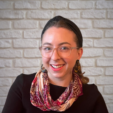 Prof. Lara Bryfonski is a white woman with blue eyes and her short brown hair in a ponytail with a headband. She is wearing large oval glasses,  a black short with a purple scarf and large earrings. 