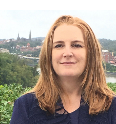 Prof. Alison Mackey is a white woman with strawberry blonde hair. She is wearing a dark blue shirt and posing in front of the Potomac River. 