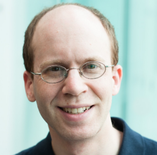 Prof. Nathan Schneider is a white man that is partially bald. He wears small rounded glasses and a dark blue collared shirt. 