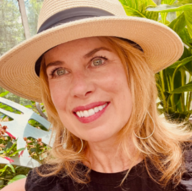 Prof. Sue Lorenson is a white woman with blonde hair and green eyes. She is wearing a sun hat and a black tank-top. 