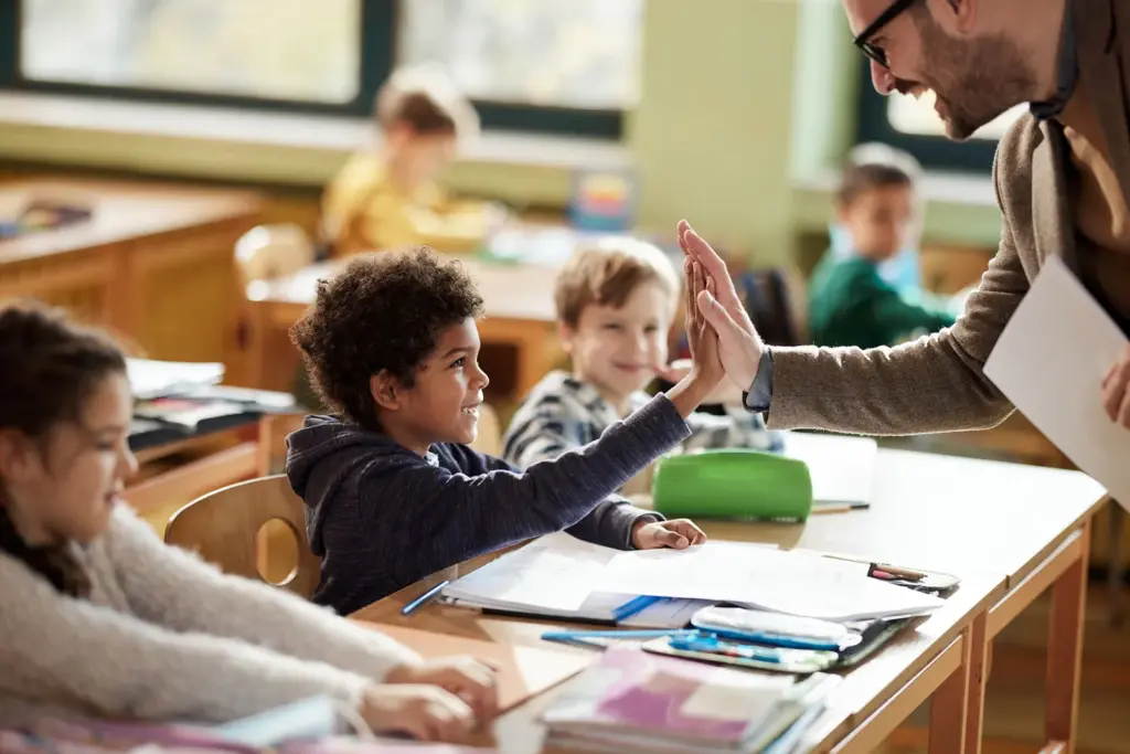 A male teacher giving a high-5 to a young boy sitting at a desk in a classroom. 