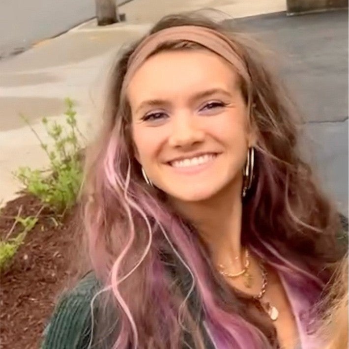 caroline gish is a white owman with long light brown hair with pink highlights. She is wearing a pink shirt with gold necklaces and large hoop earrings. 