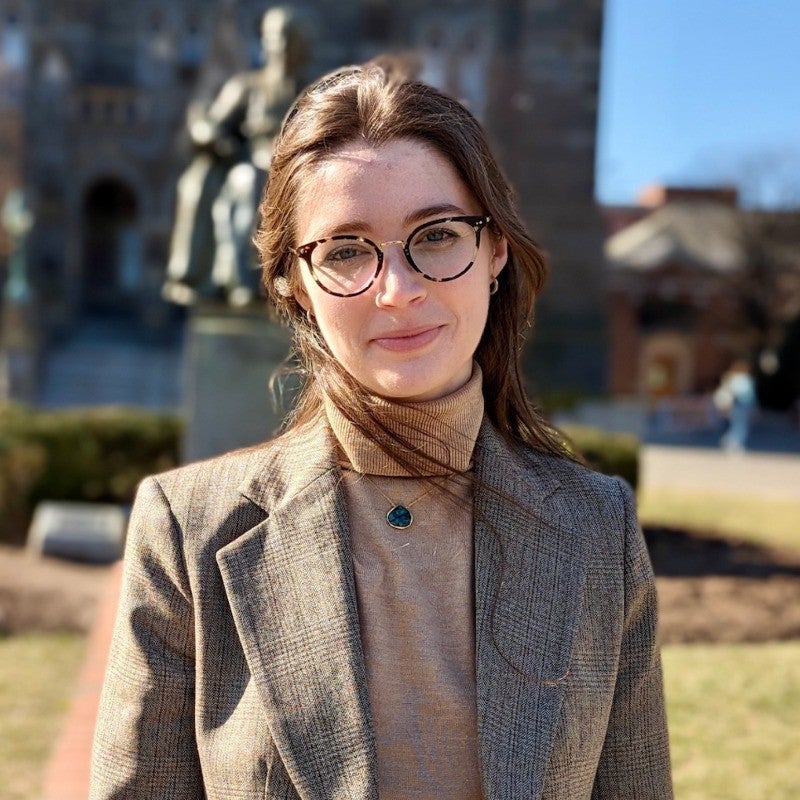 Erin as a white woman with brown hair and large glasses. She is wearing a dark beige shirt and blazer. 