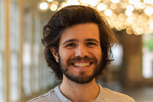 This is a portrait of Prof. Ethan Gotlieb Wilcox, who is a young white man with medium length dark brown hair and a beard. He is wearing a grey t-shirt and a large smile, 