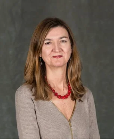 Prof. Lourdes Ortega is a Latina woman with medium-length light brown hair and brown eyes. She is wearing a grey blouse with a red beaded necklace. 