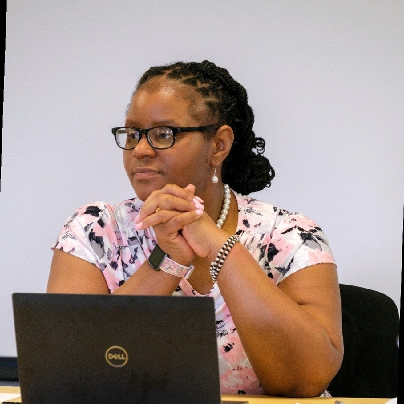 Minnie is a black woman with braid pulled back into a ponytail and black squared glasses. She is listening with a laptop in front of her. 