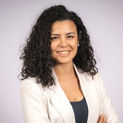 Seyma is a woman with curly black hair, wearing a white blazer and black top. 