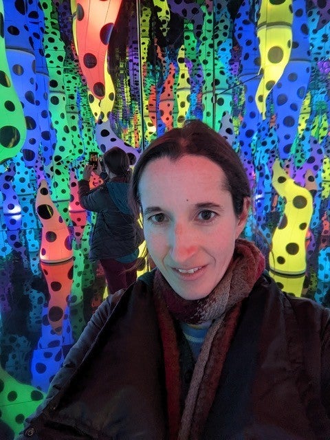 Francesca Di Silvio smiling in a colorful dotted light installation, wearing a scarf and coat
